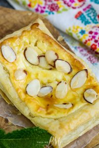 An Almond Puff Pastry Danish graces the wooden board, its surface adorned with delicate sliced almonds, while a vibrant fabric backdrop adds a burst of color to the scene.