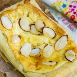 An Almond Puff Pastry Danish graces the wooden board, its surface adorned with delicate sliced almonds, while a vibrant fabric backdrop adds a burst of color to the scene.