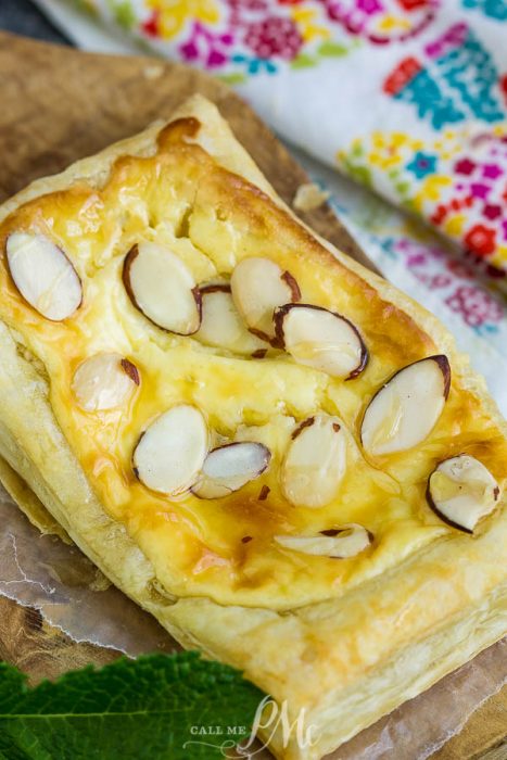 An Almond Puff Pastry Danish graces the wooden board, its surface adorned with delicate sliced almonds, while a vibrant fabric backdrop adds a burst of color to the scene.