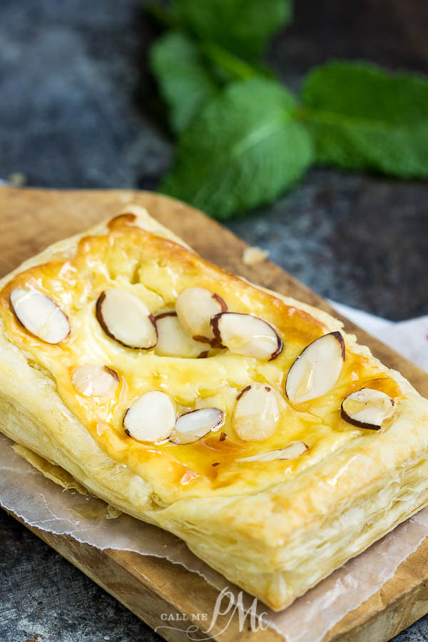 Almond Puff Pastry Danish takes advantage of premade puff pastry for the no-fail pastry base that's filled with a cream cheese honey mixture & topped with almond slivers. #puffpastry #danish #recipe #breakfast #brunch #dessert #almond #baked #homemade #semihomemade A dessert graces the wooden board, its surface adorned with delicate sliced almonds, while a vibrant fabric backdrop adds a burst of color to the scene.