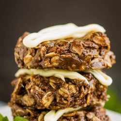 A stack of three healthy no-bake oatmeal chocolate cookies topped with white icing, placed on a pristine surface with a mint leaf beside them.