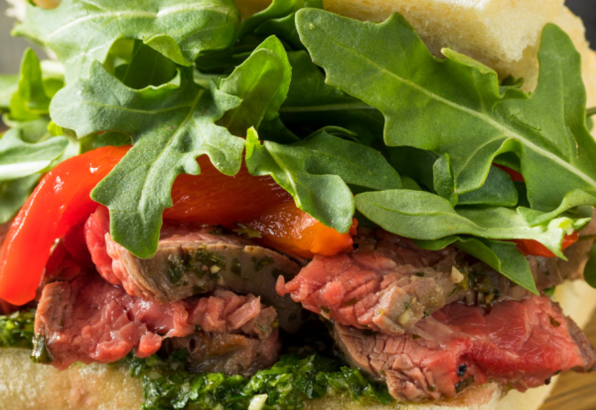 Close-up of a meal with slices of rare meat, red bell pepper, pesto, and fresh arugula on a crusty bread roll.