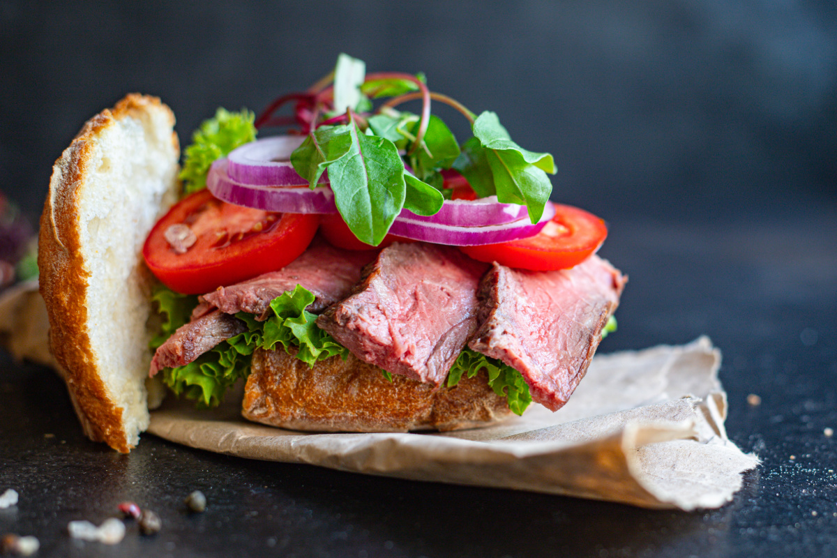Open-faced steak sandwich with slices of roast beef, tomato, red onion, leafy greens, and lettuce on crusty bread, placed on parchment paper against a dark background.