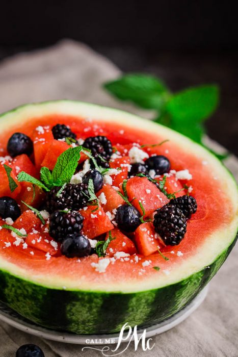 Half of a melon serves as a bowl for this vibrant dish, featuring blackberries, blueberries, crumbled white cheese, and fresh mint leaves on a beige cloth with a dark background.