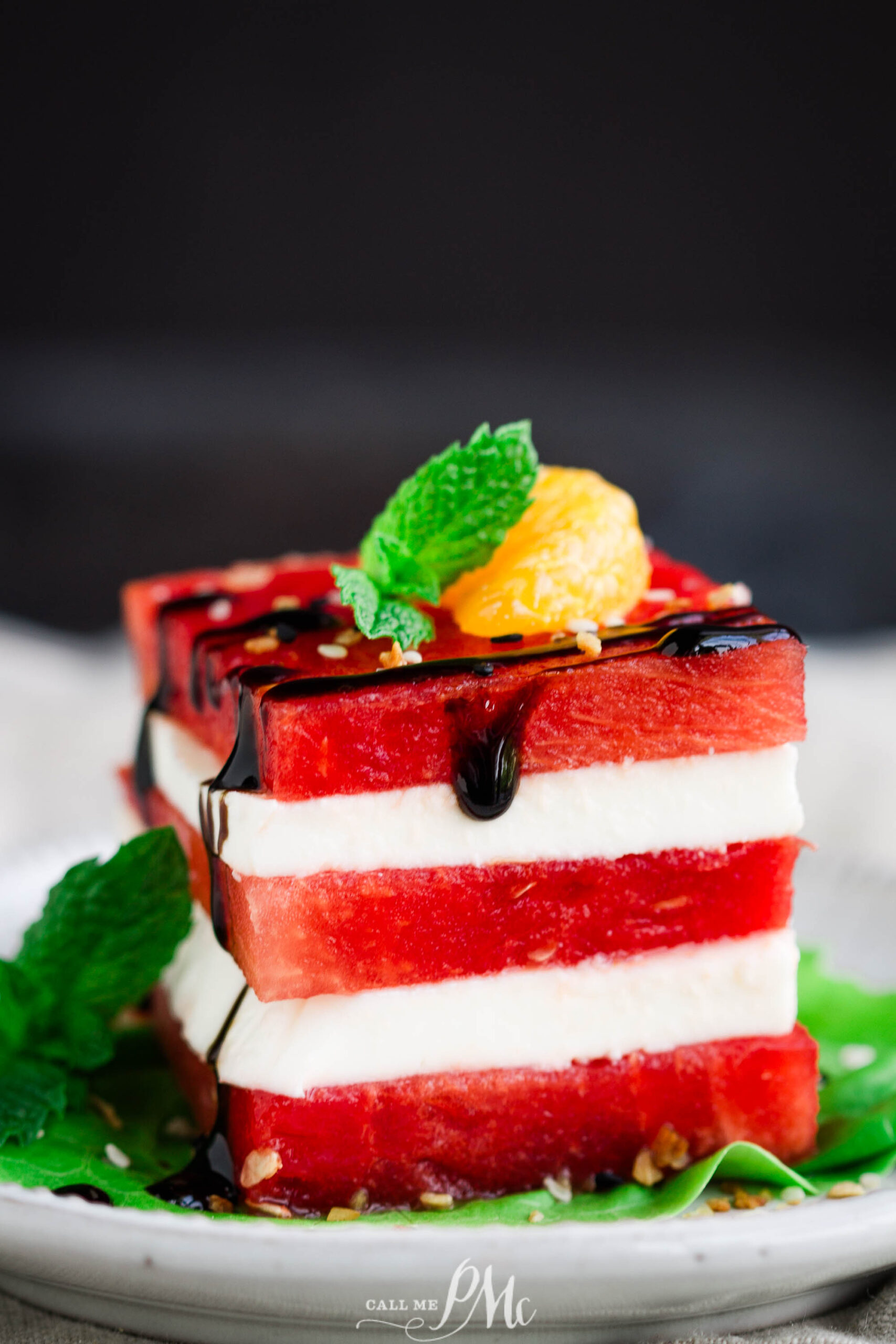 A refreshing Watermelon Salad featuring alternating slices of watermelon and feta cheese, garnished with a mint sprig, a small yellow fruit, and a drizzle of balsamic glaze, served on a white plate.