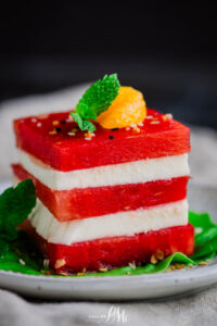 A vibrant Watermelon Salad features a stack of alternating watermelon and white cheese slices, garnished with mint leaves, an orange segment, and sesame seeds, all elegantly arranged on a white plate.