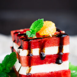 A refreshing Watermelon Salad features stacked watermelon and white cheese slices, garnished with a mint sprig, a small yellow fruit, and drizzled with balsamic glaze on a white plate.