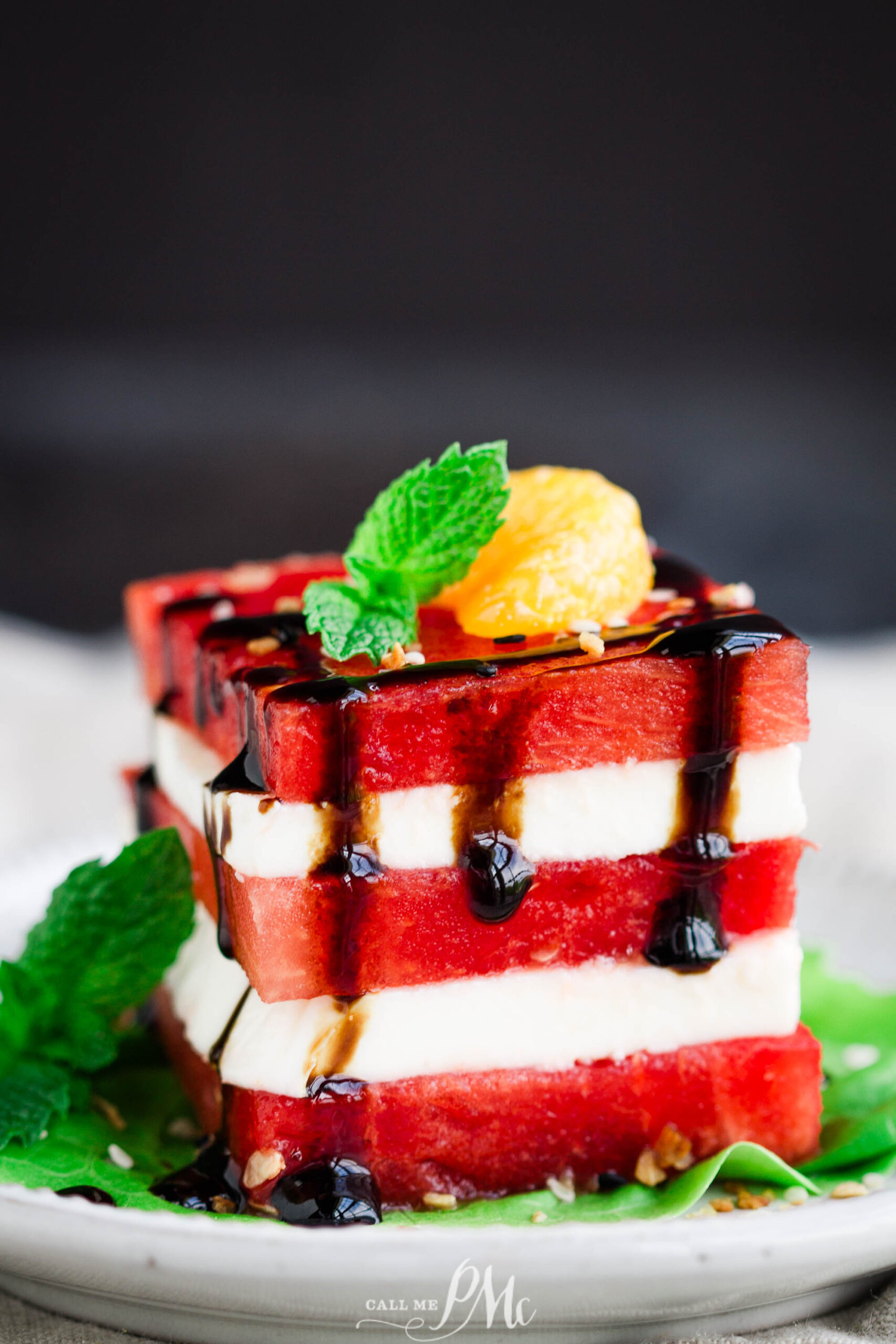 A refreshing Watermelon Salad features stacked watermelon and white cheese slices, garnished with a mint sprig, a small yellow fruit, and drizzled with balsamic glaze on a white plate.