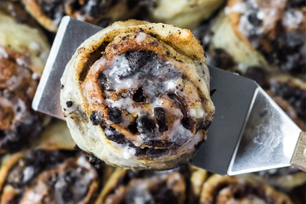 Oreo cinnamon bun on a metal spatula above a pan full of them.