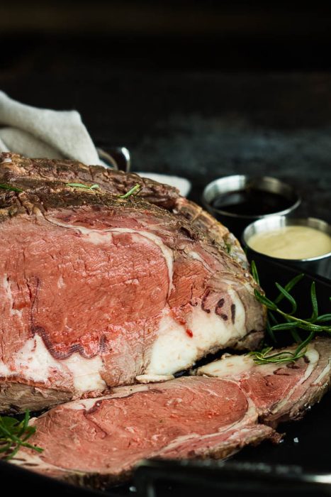 A sliced Boneless Prime Rib Roast on a dark surface, garnished with rosemary, with small bowls of sauce in the background.