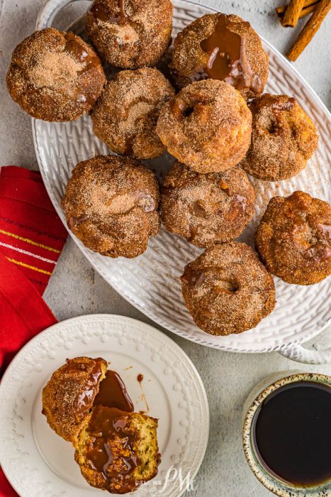 Churro Muffins A white platter holds several cinnamon sugar-coated donuts, some drizzled with chocolate sauce. Nearby, Dulce de Leche Churro Muffins sit beside a halved chocolate-filled donut, a cup of coffee, and a red napkin.