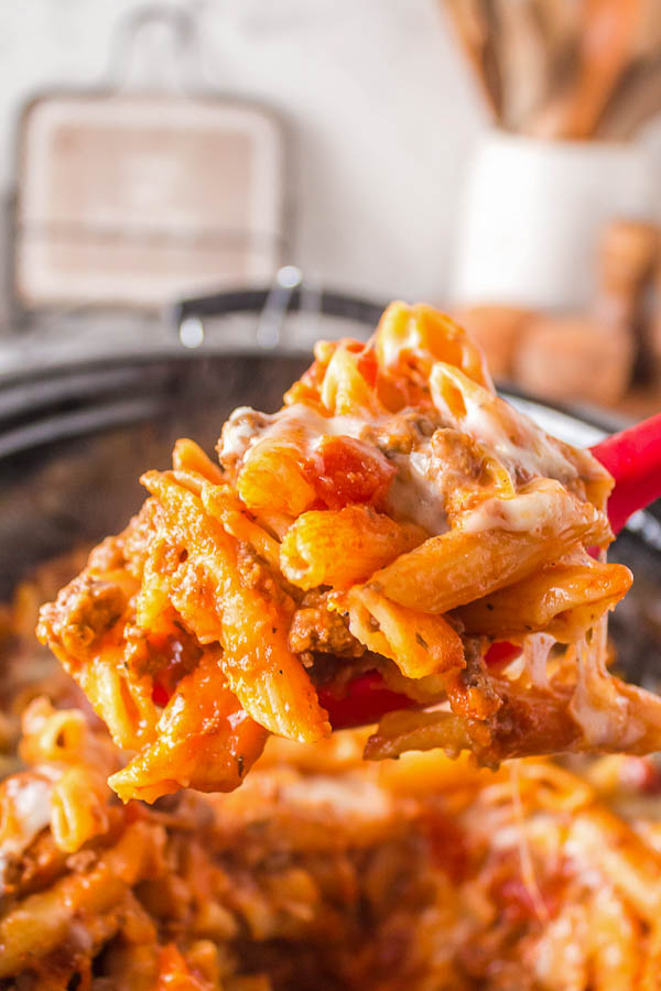 The jam-packed crock pot is being emptied of some delightful cheesy penne using a red spoon.