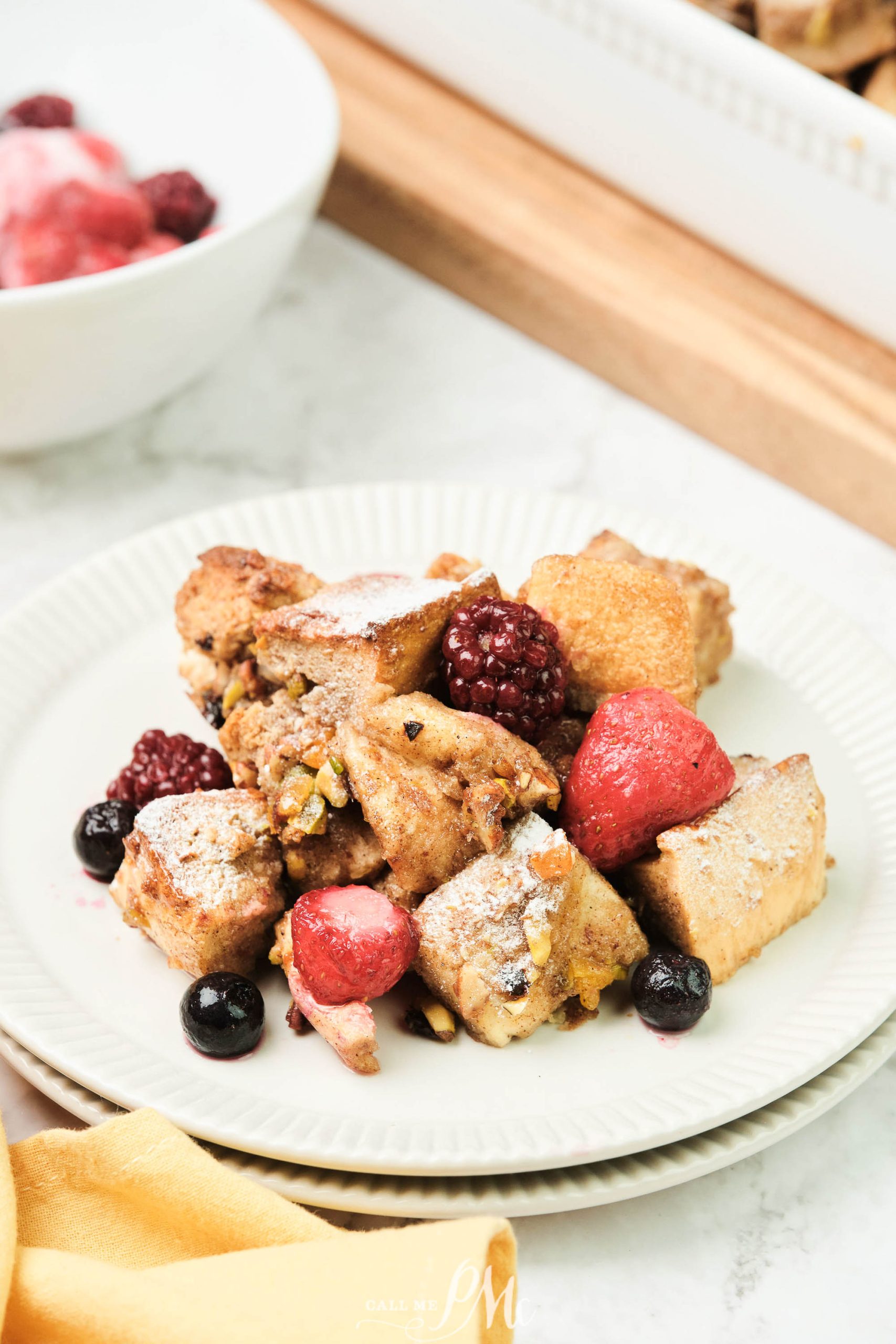 A plate of dessert is topped with strawberries, blackberries, and blueberries, dusted with powdered sugar. A bowl with more berries is in the background.