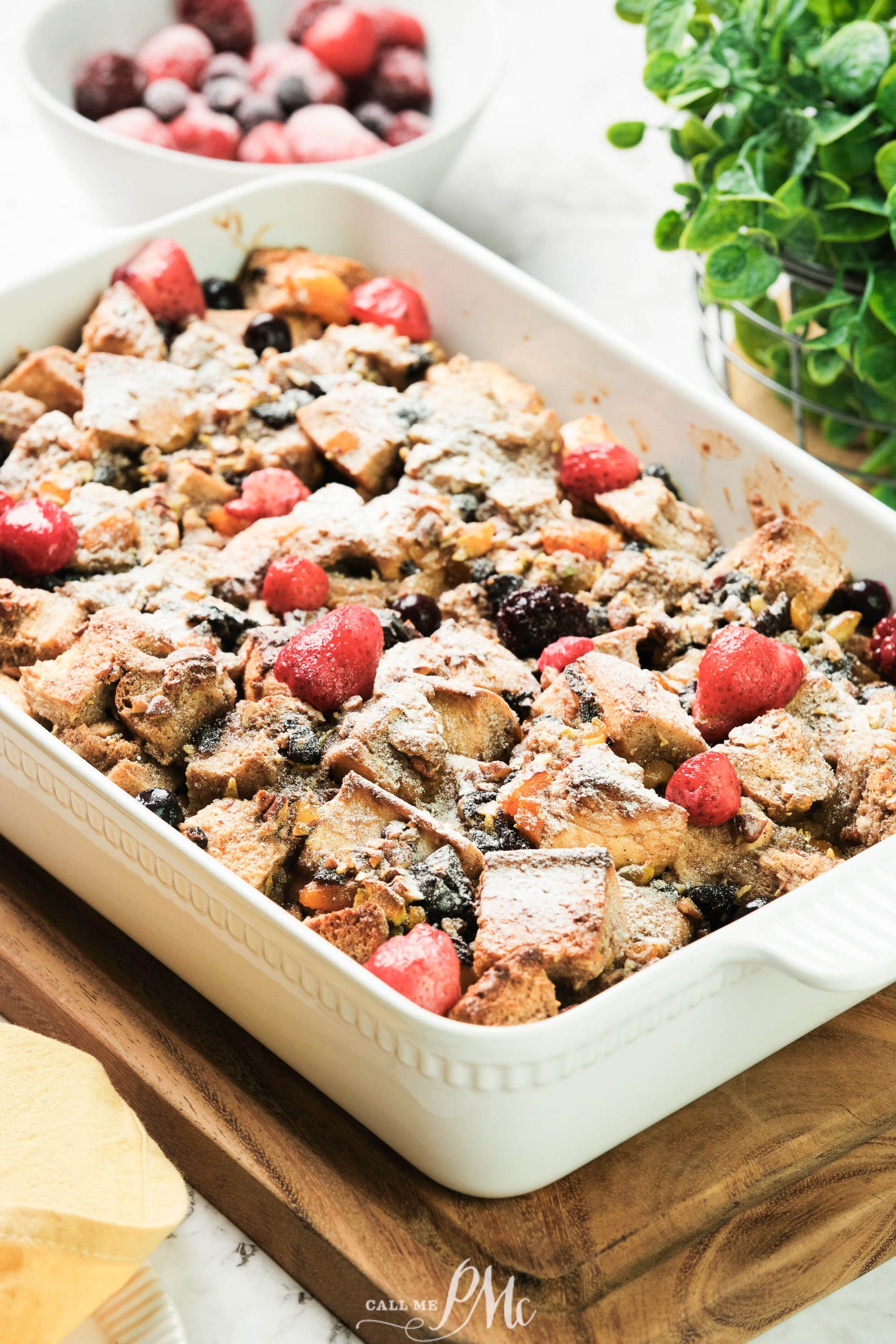 A plate of dessert is topped with strawberries, blackberries, and blueberries, dusted with powdered sugar. A bowl with more berries is in the background.