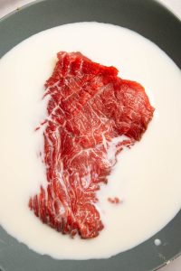 Raw cube steak in a bowl with milk covering part of it.