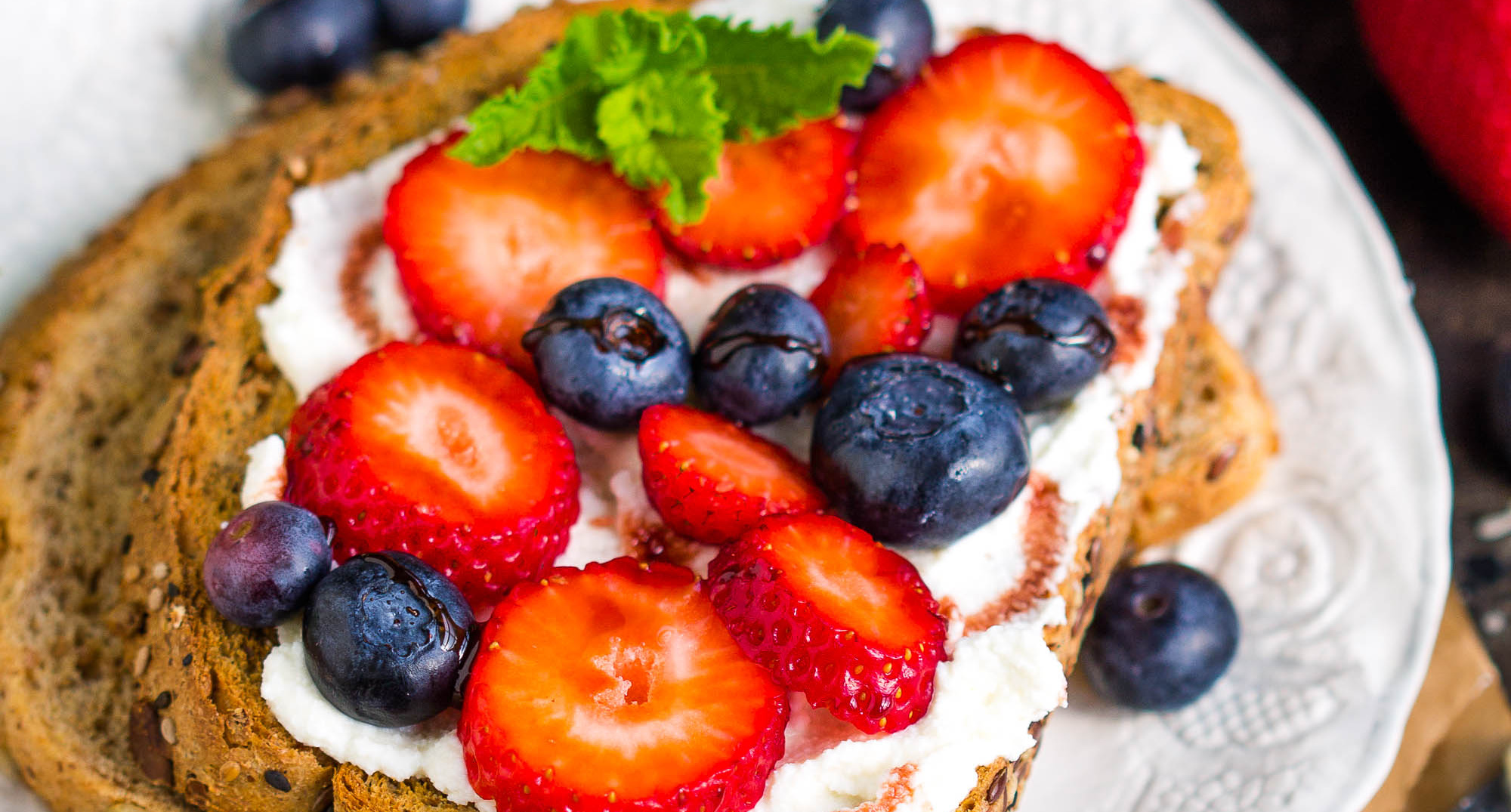 Date Syrup Drizzled Berry Ricotta Toast: A slice of multigrain bread topped with ricotta cheese, sliced strawberries, blueberries, a sprig of mint, and finished with a drizzle of date syrup on a white plate.