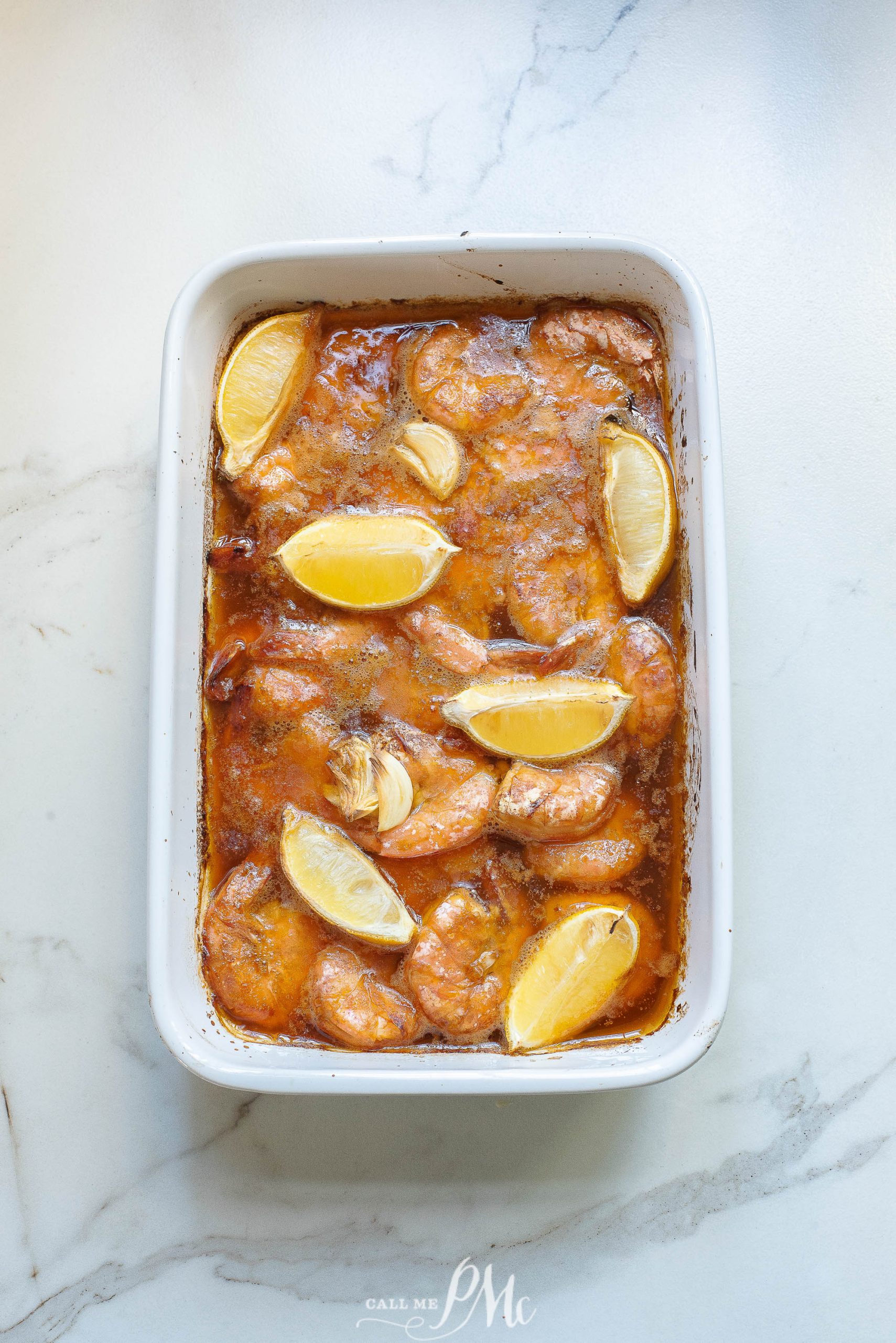 Shrimp with lemon wedges in a red sauce in a white casserole dish.