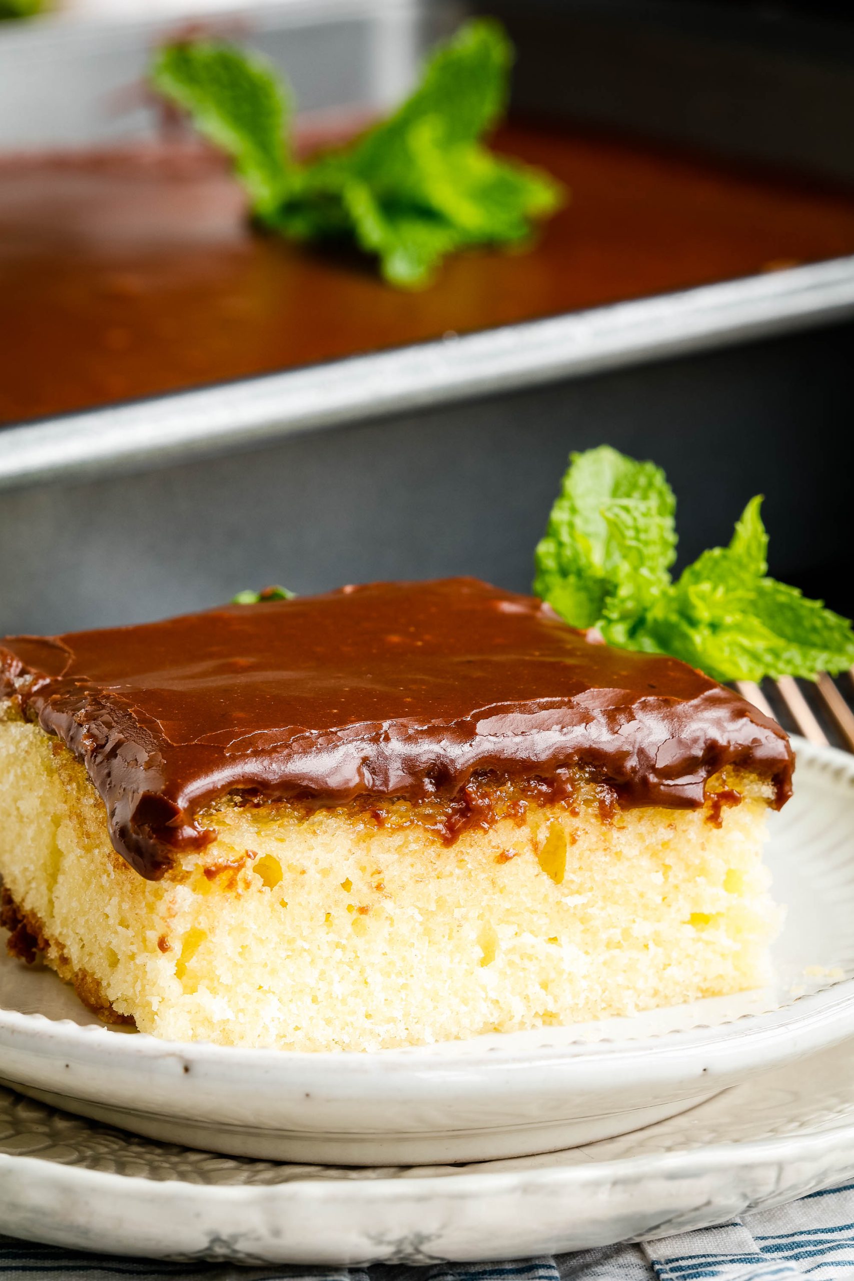 Square piece of vanilla cake with chocolate frosting on a white plate.