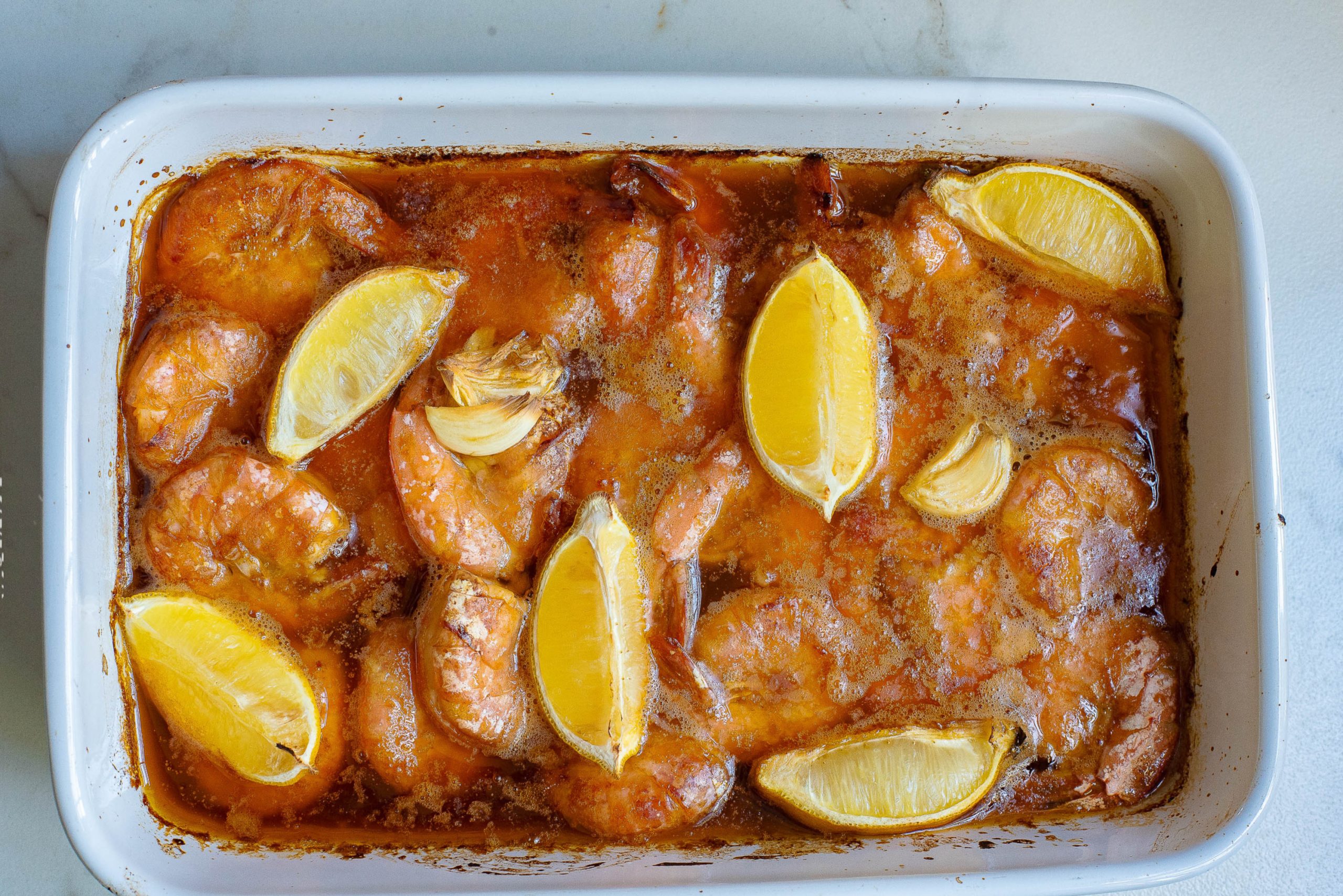 Shrimp with lemon wedges in a red sauce in a white casserole dish.