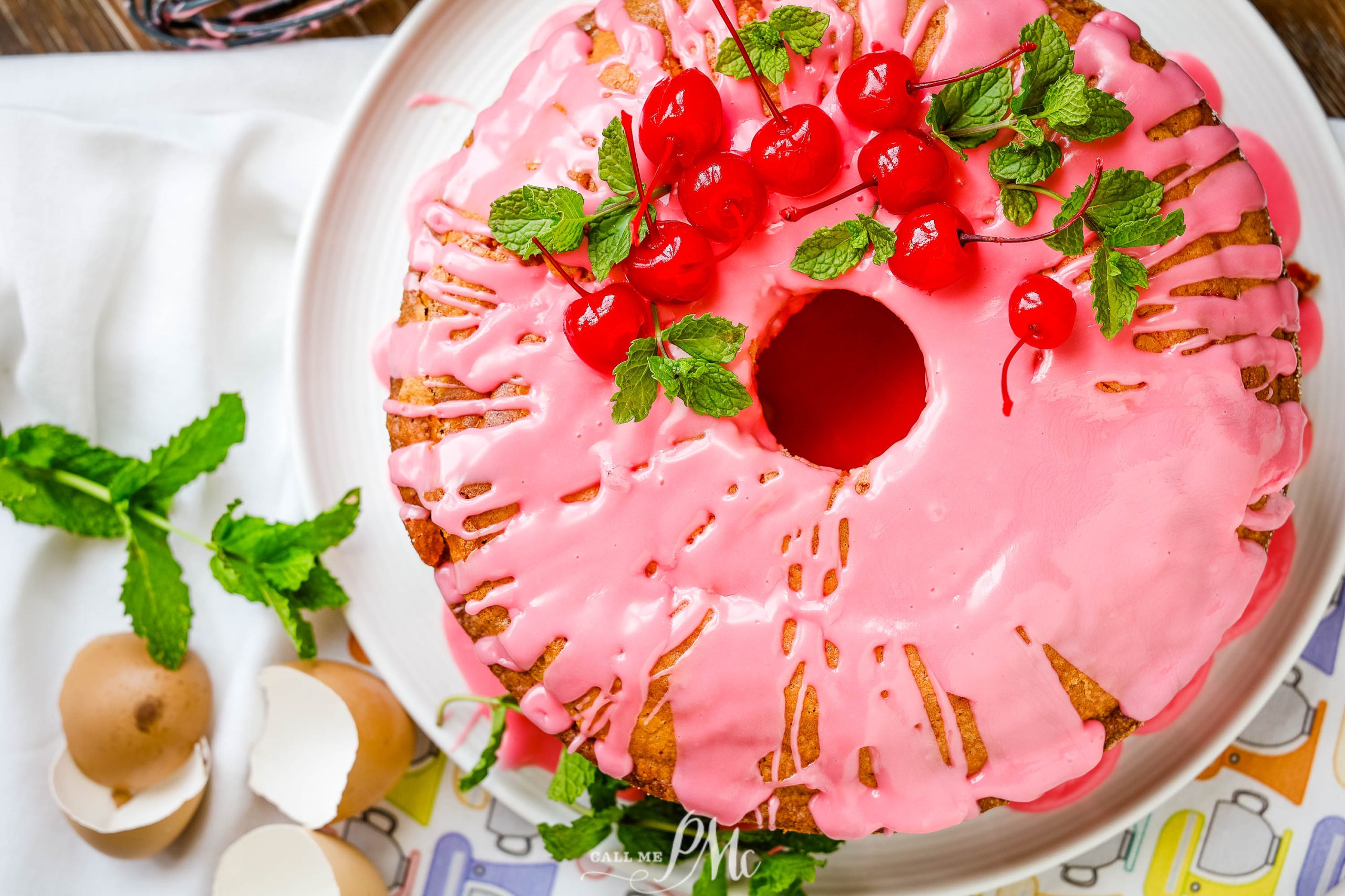 Flatlay of whole pound cake with pink frosting and cherries.