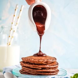 A person pouring chocolate sauce on a stack of pancakes.