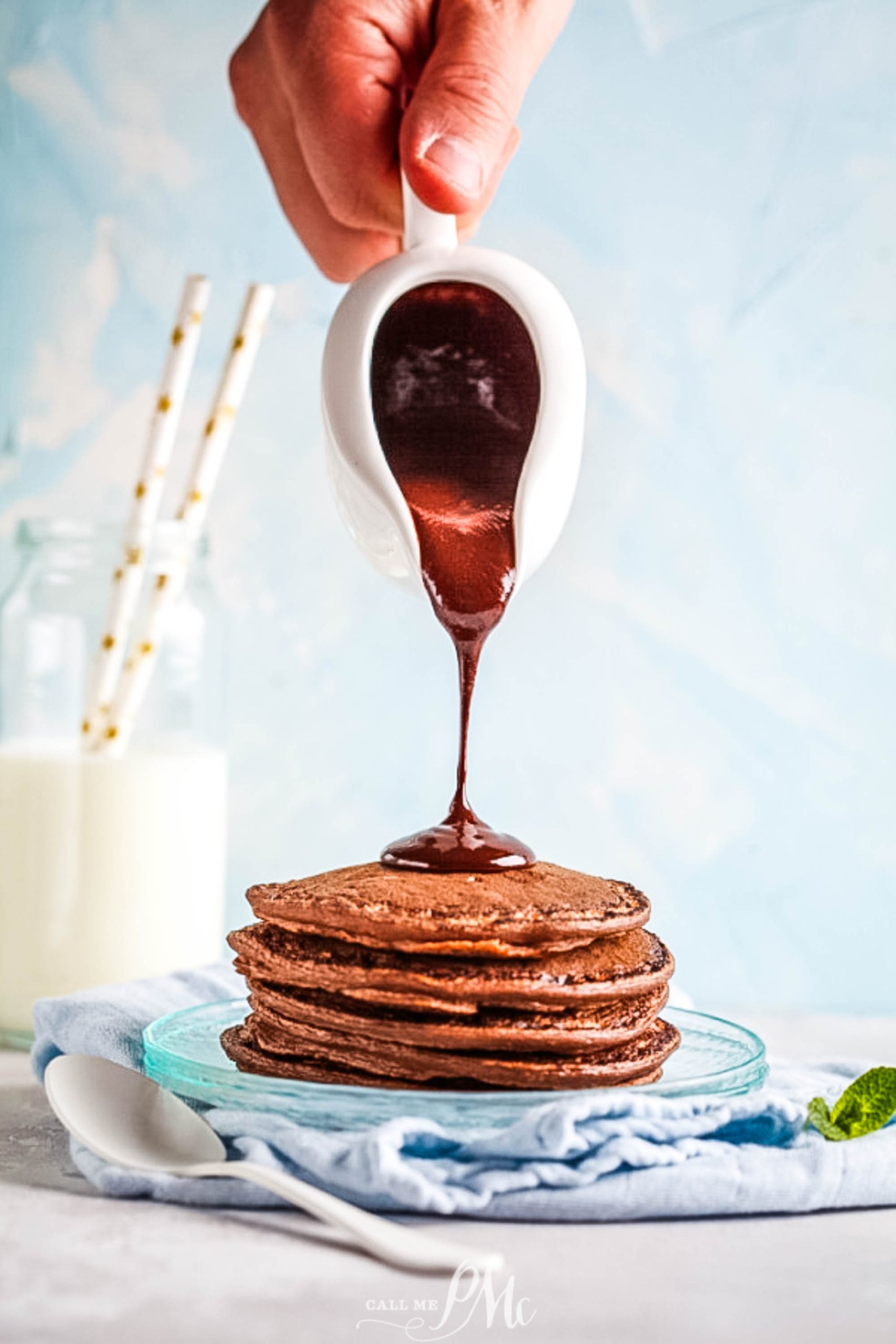 A person pouring chocolate sauce on a stack of pancakes.