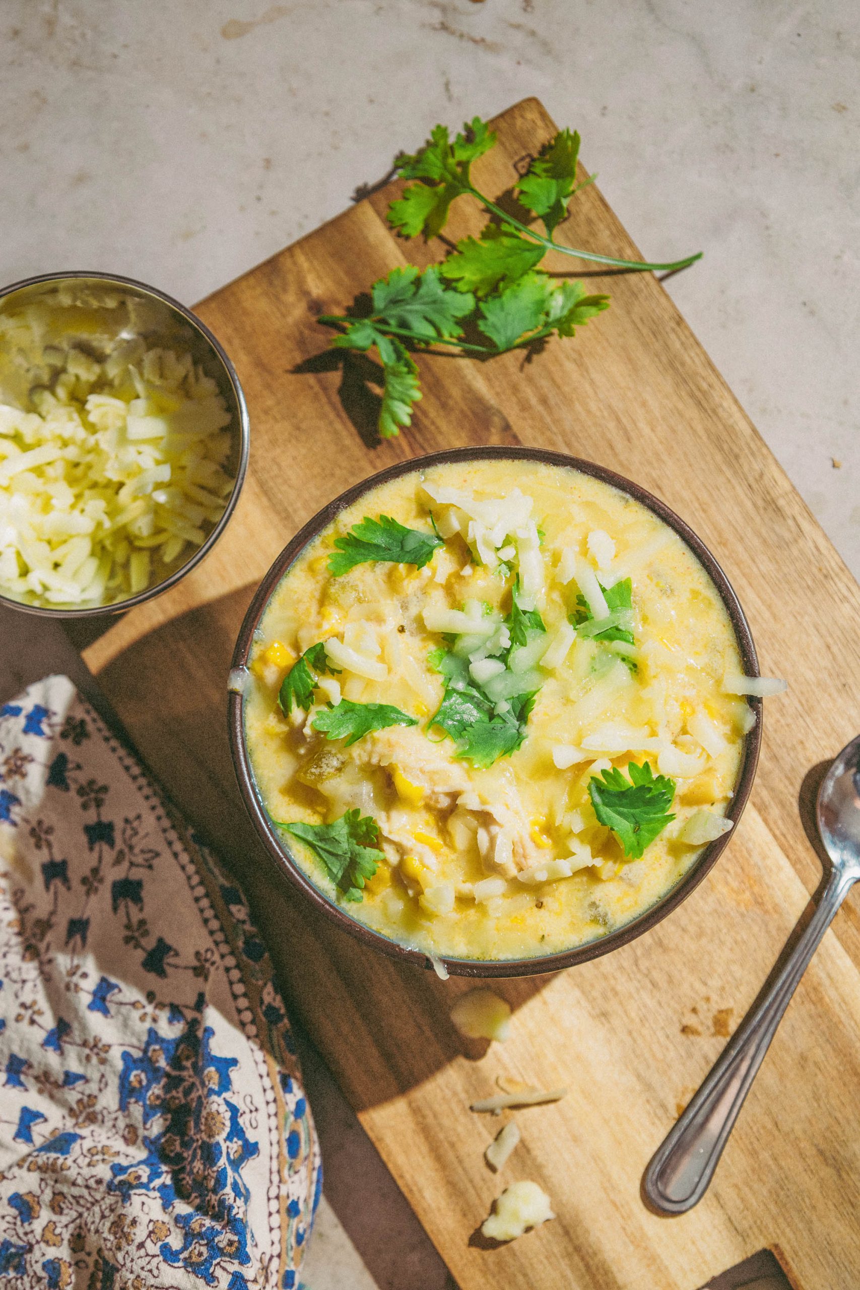 A bowl of Green Chili Chicken Corn Chowder and a spoon on a wooden cutting board.