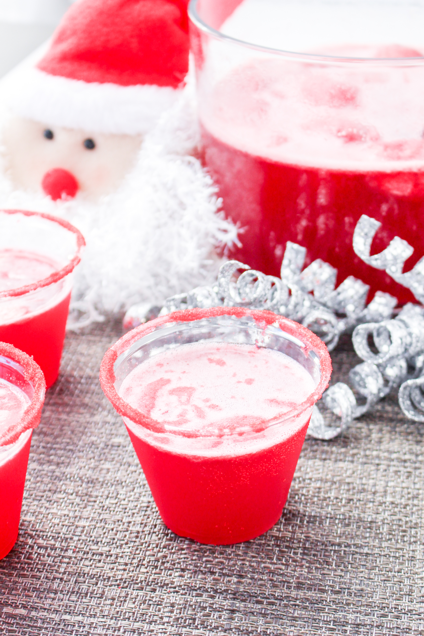 Santa claus is sitting on a table next to cups of red liquid.