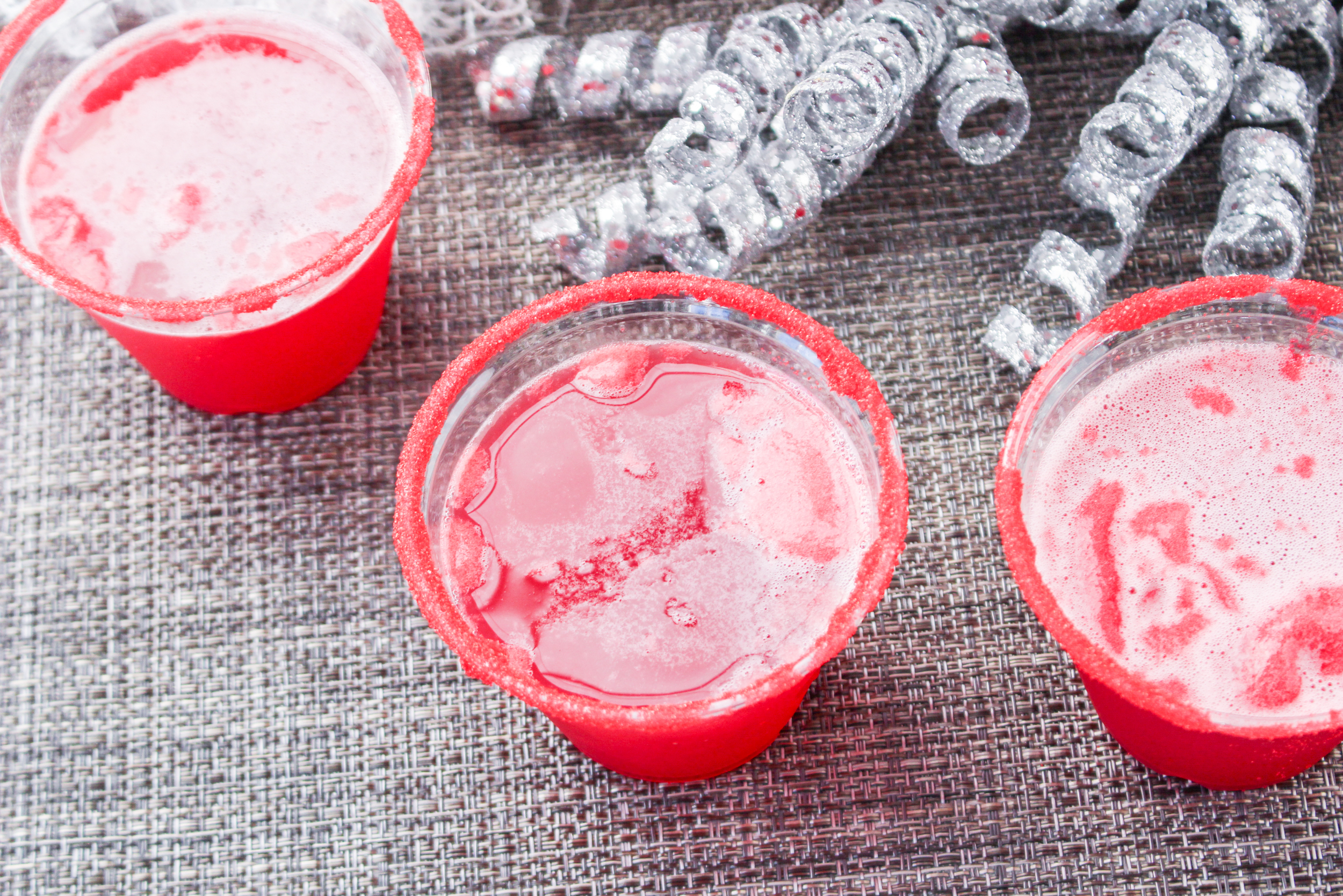 Three cups of red liquid on a table with silver confetti.