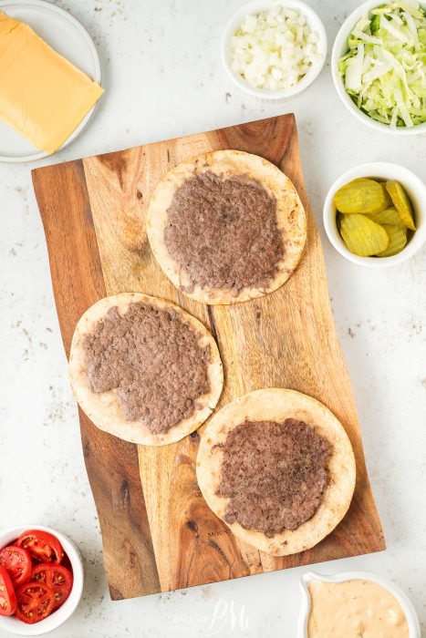 A wooden cutting board with burgers and toppings on it.