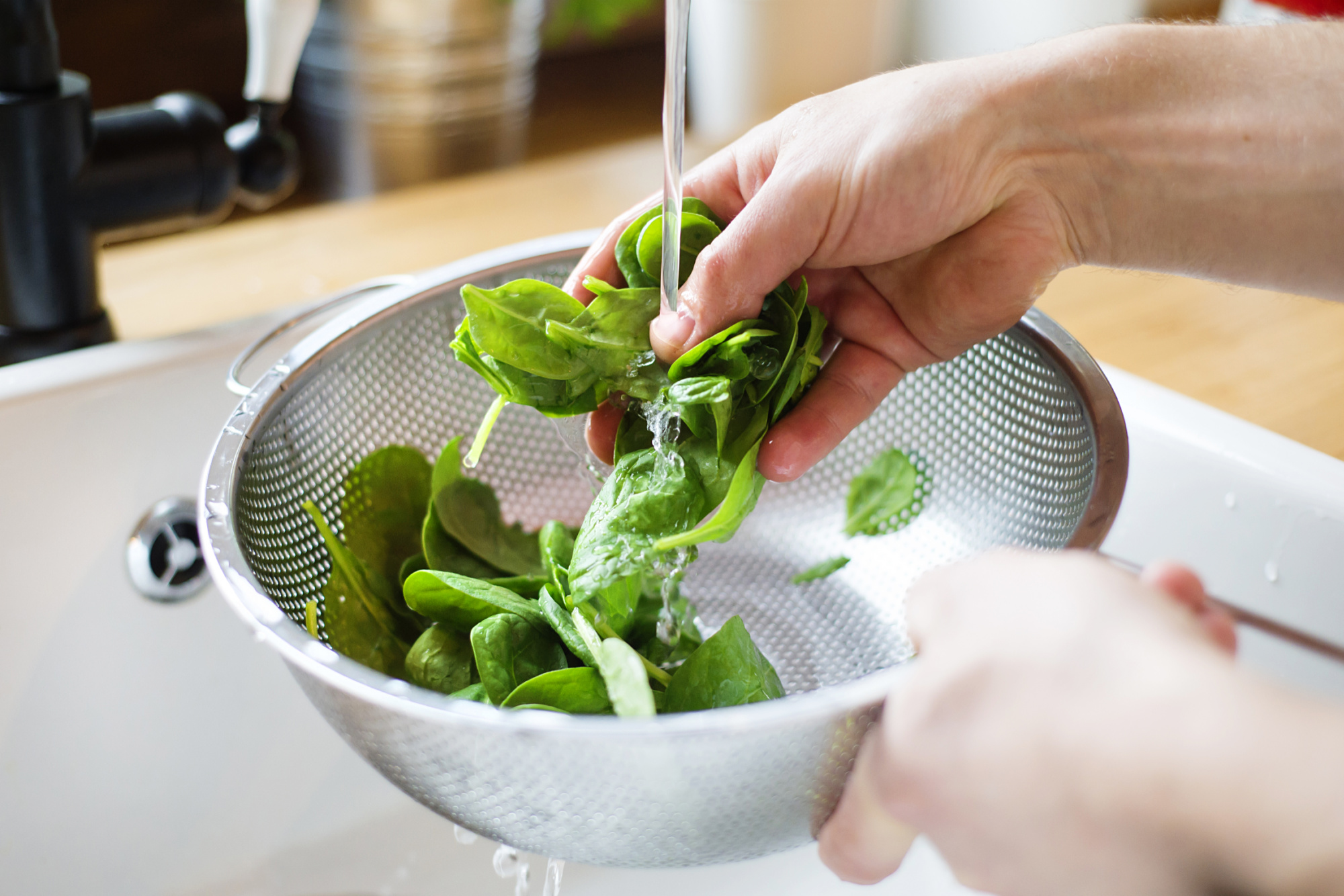 A person is rinsing organic spinach in a colander using vegetable wash.