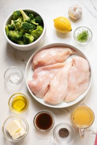 Chicken breasts, broccoli, lemon, and other ingredients on a marble table.