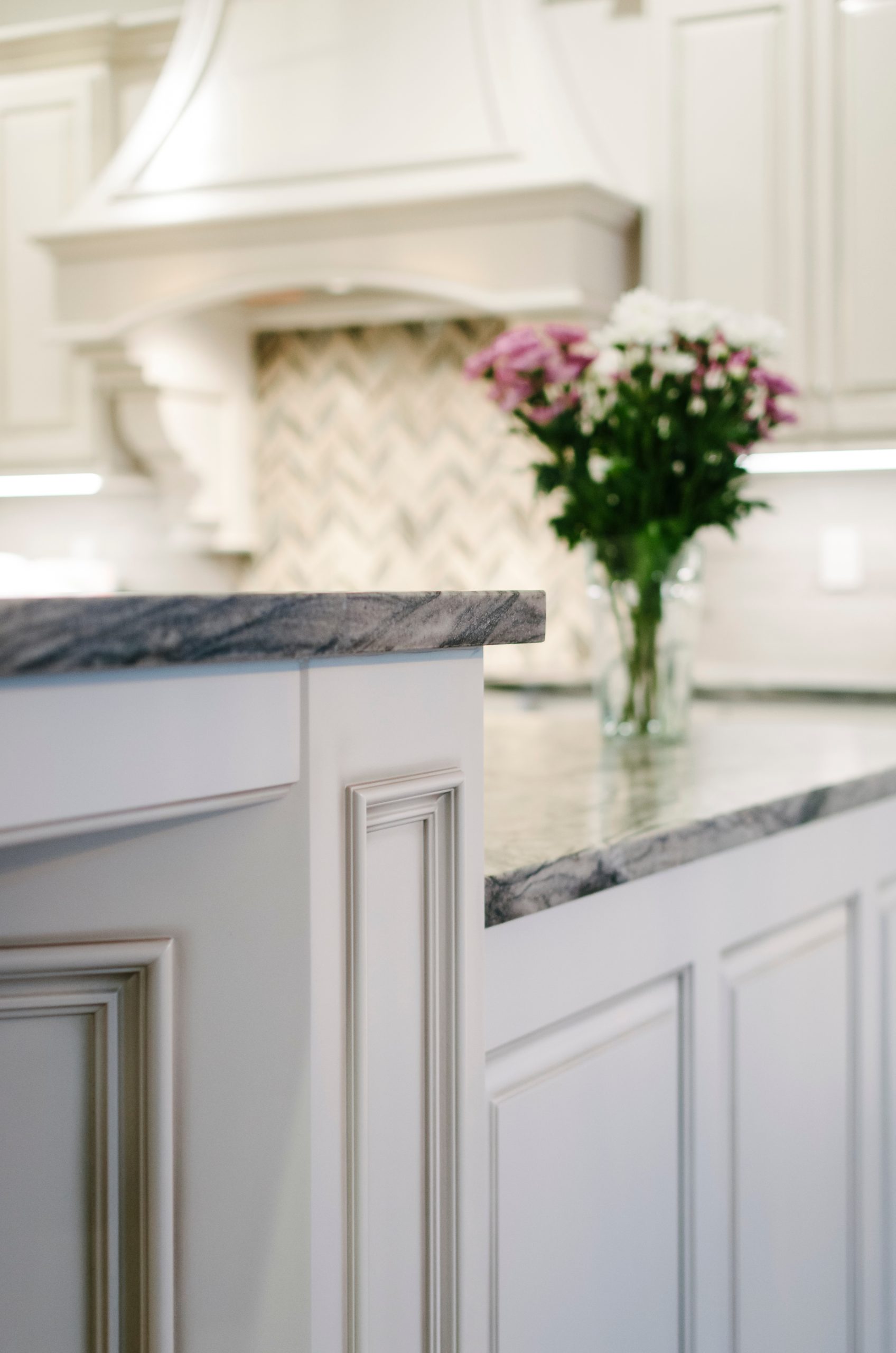 A white kitchen with marble counter tops and flowers.