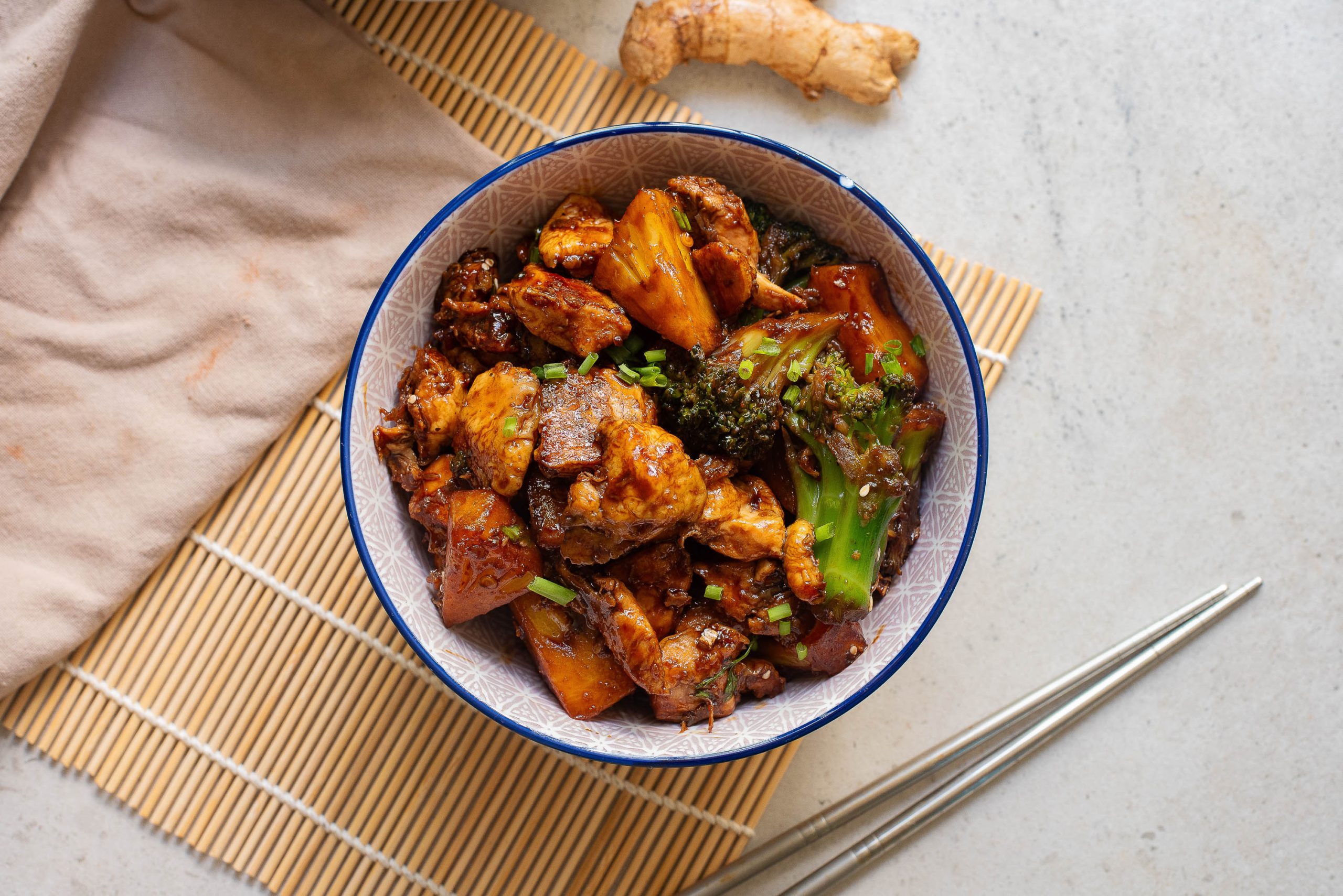 A bowl of chicken and broccoli with chopsticks on a mat.