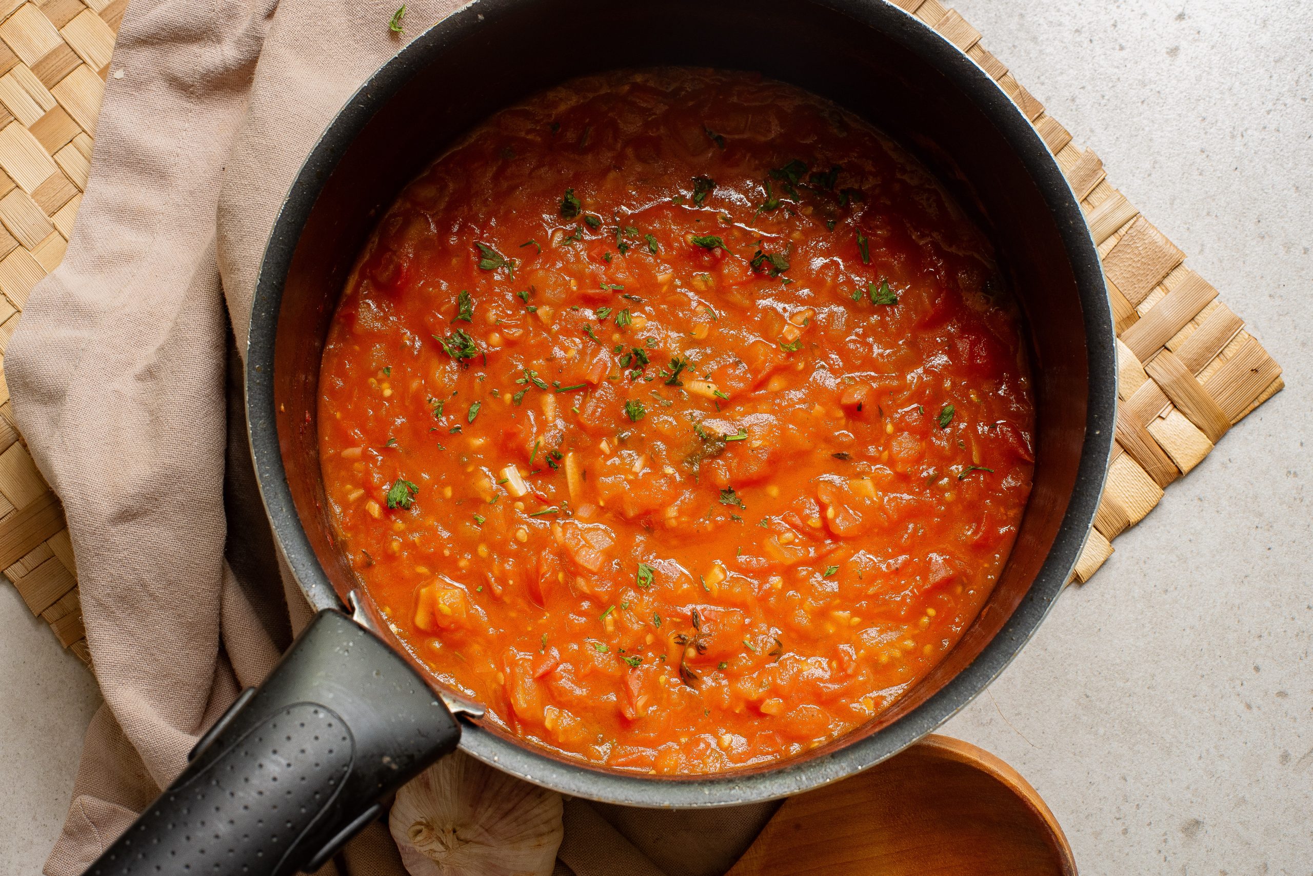 A pot of Simple Homemade Marinara Saucewith herbs, accompanied by a wooden spoon, on a kitchen counter.