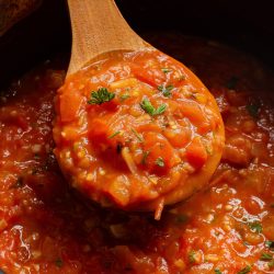 A wooden spoon scooping tomato sauce with vegetables from a pot.