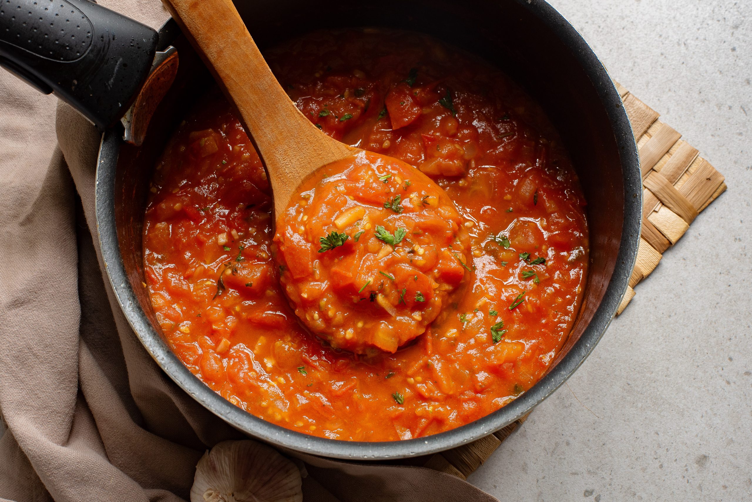 A pot of Simple Homemade Marinara Saucewith herbs, accompanied by a wooden spoon, on a kitchen counter.