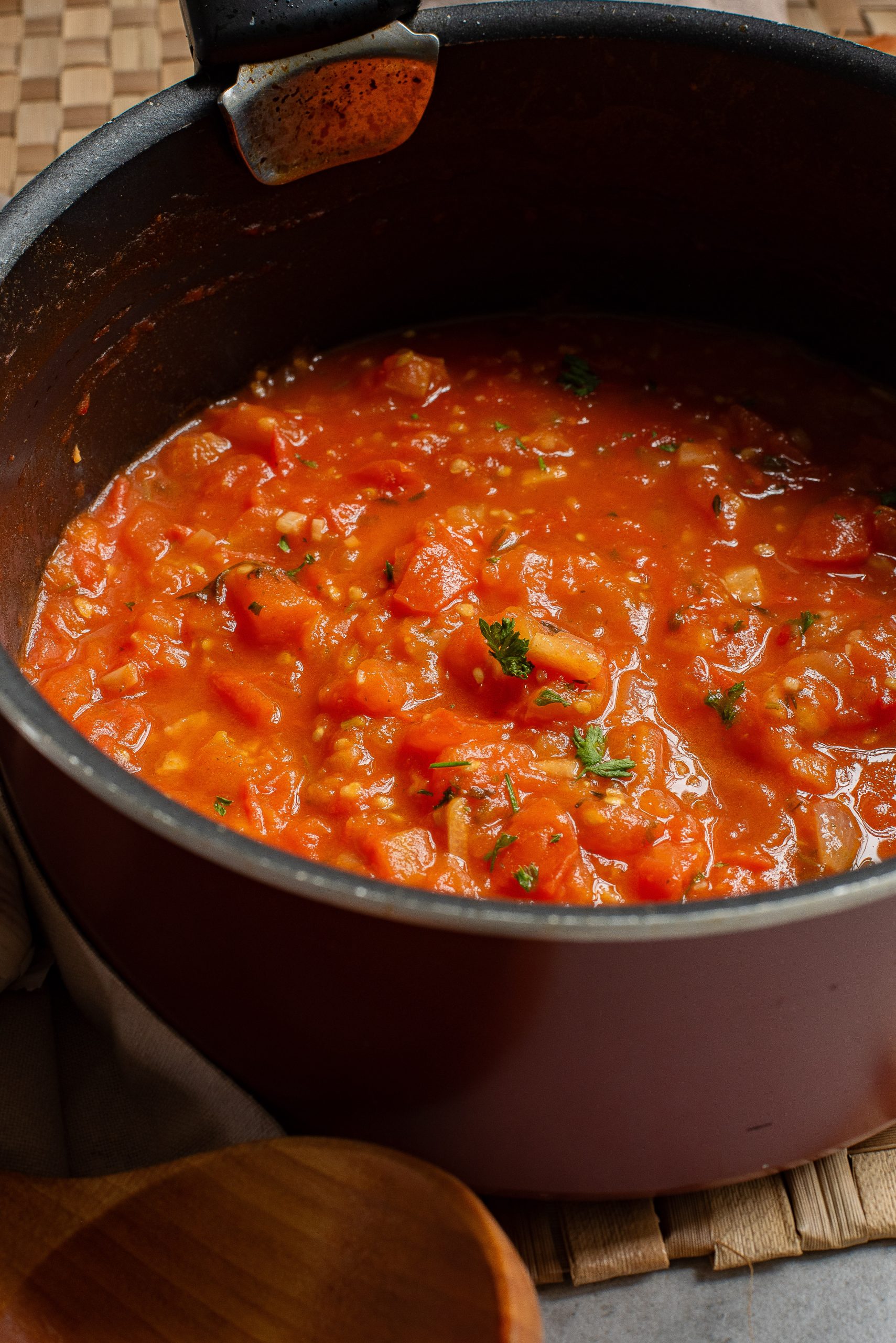 A pot of freshly cooked Simple Homemade Marinara Sauce garnished with herbs, with a wooden spoon on the side.