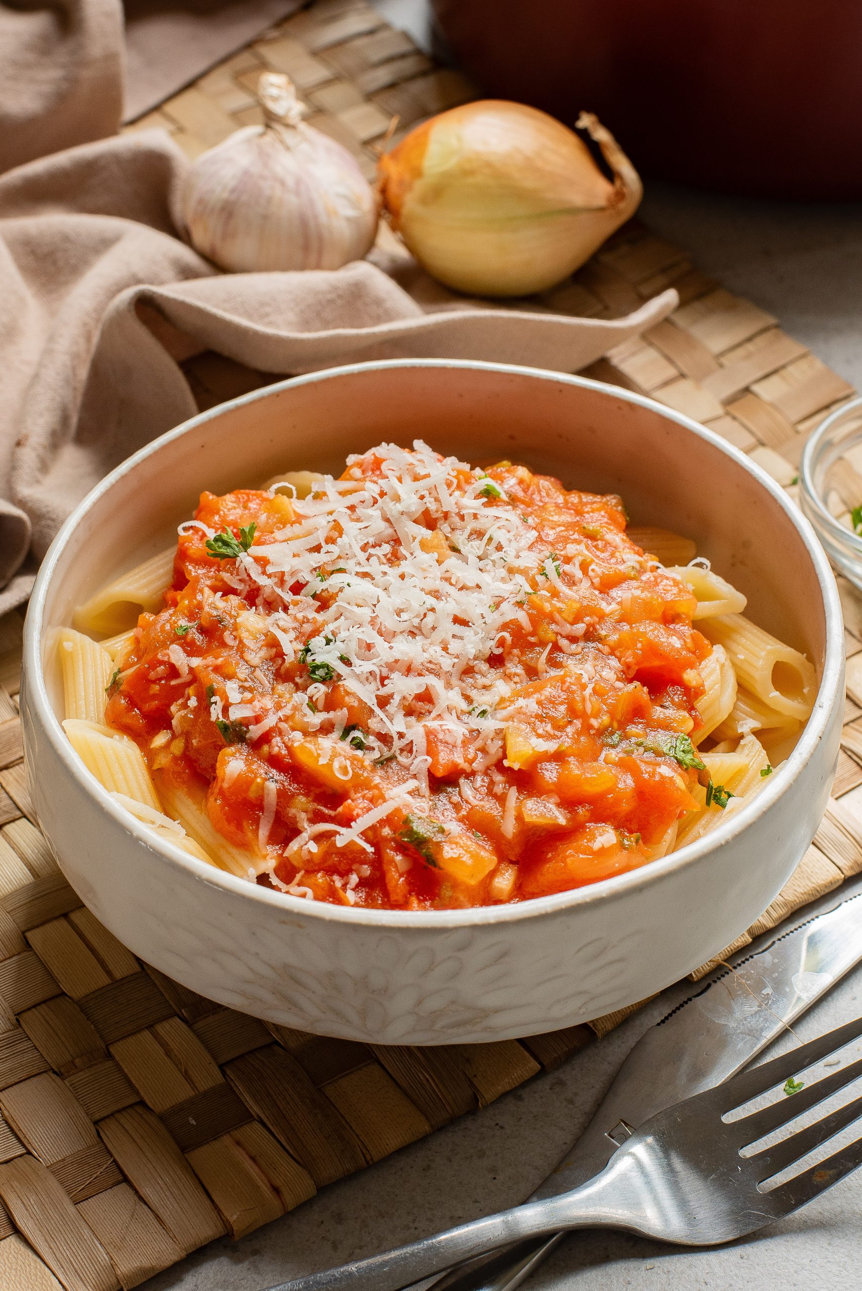 A bowl of pasta with tomato sauce and grated cheese, served on a woven placemat with a fork on the side.