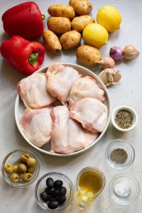 Ingredients for cooking arrayed on a surface, including raw chicken thighs, vegetables, lemons, herbs, and olives.