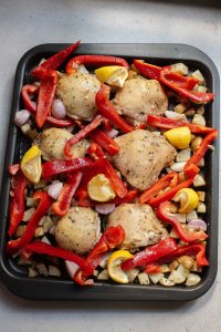 A tray of uncooked chicken thighs with red bell peppers, lemon slices, and garlic cloves prepped for roasting.