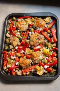Roasted chicken thighs with bell peppers, olives, and garlic on a baking sheet.