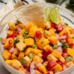 A bowl of mango salsa with a lime wedge and tortilla chip on the side.