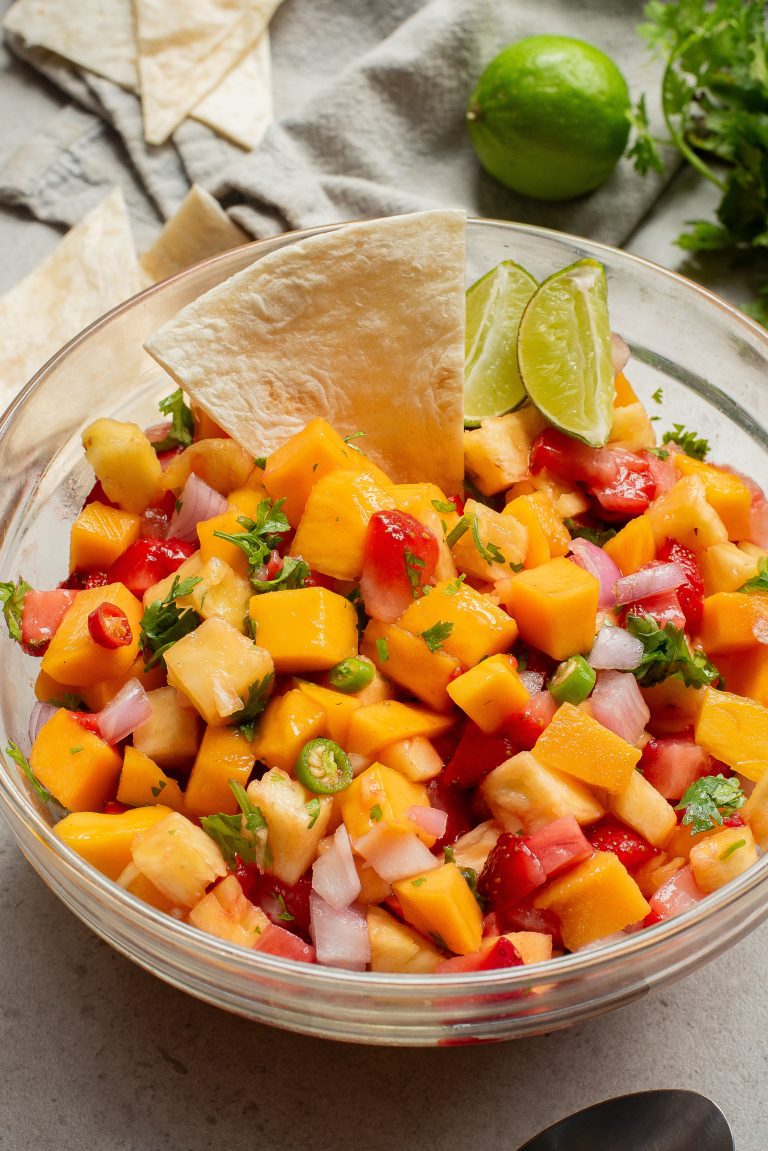 A bowl of mango salsa with a lime wedge and tortilla chip on the side.