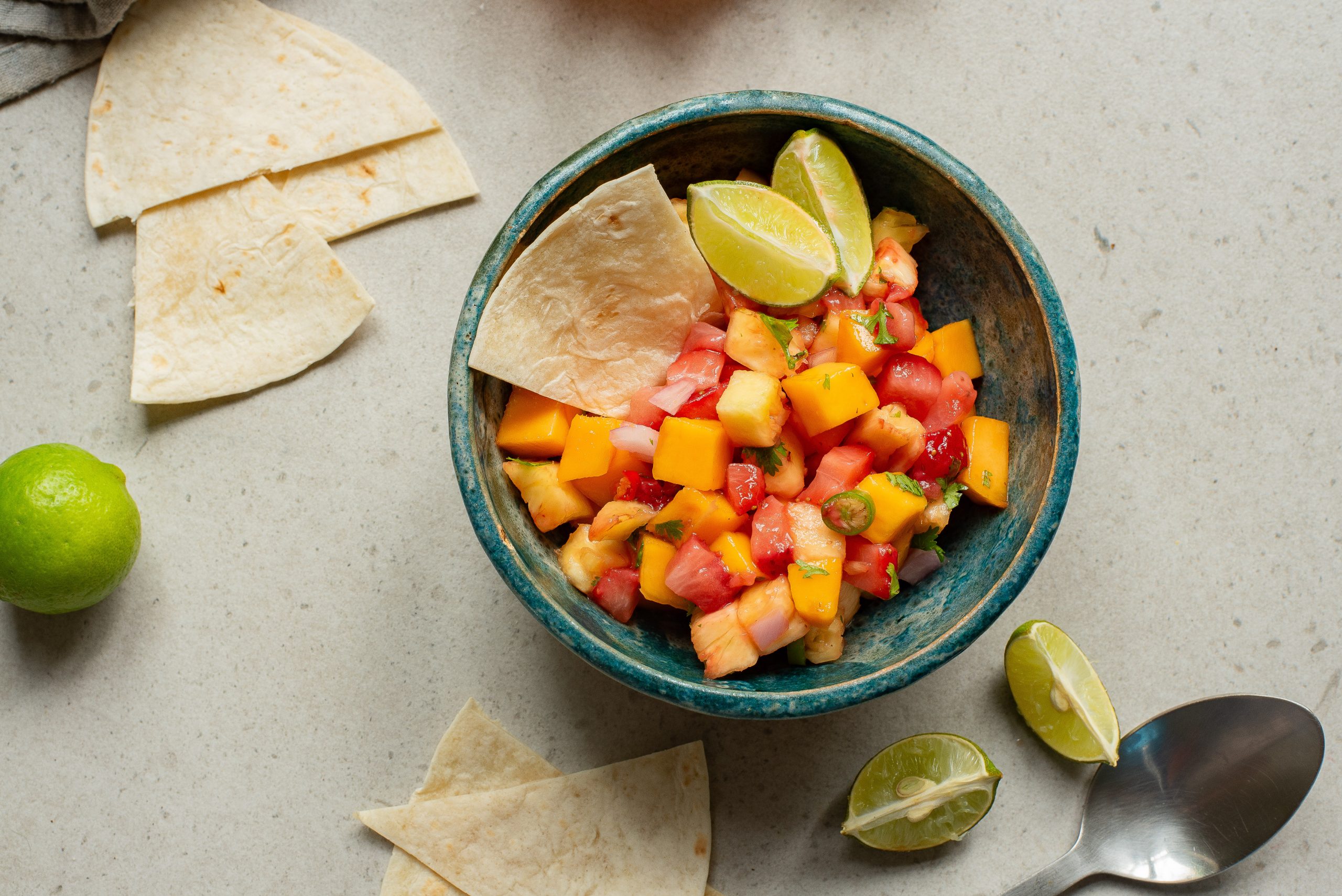 A bowl of fresh fruit salsa with diced mango, tomato, and lime, accompanied by tortilla wedges, on a neutral-toned surface.