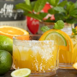 Glass of citrus cocktail garnished with orange slice and mint, surrounded by fresh fruits and bottles.