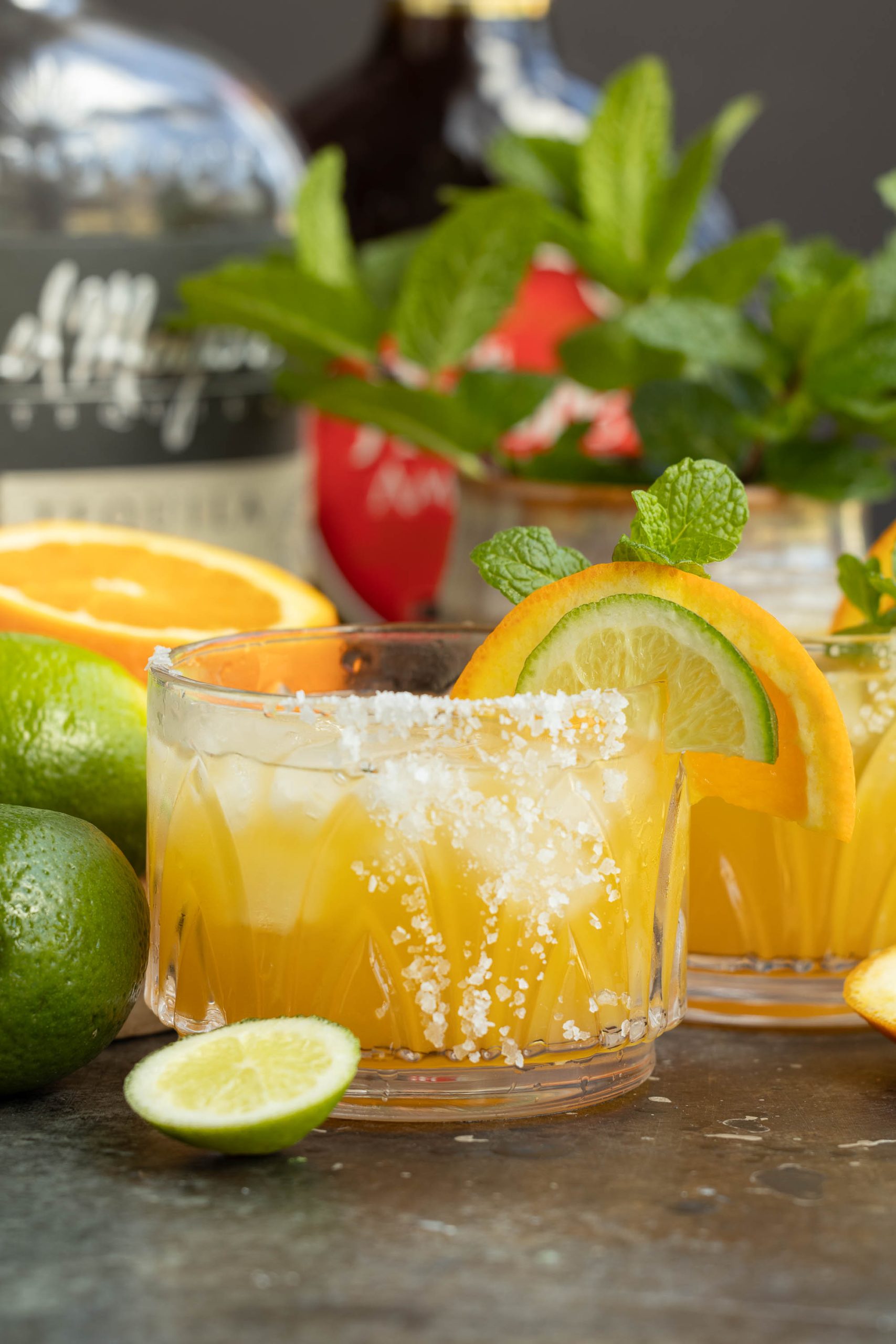 Glass of citrus cocktail garnished with orange slice and mint, surrounded by fresh fruits and bottles.