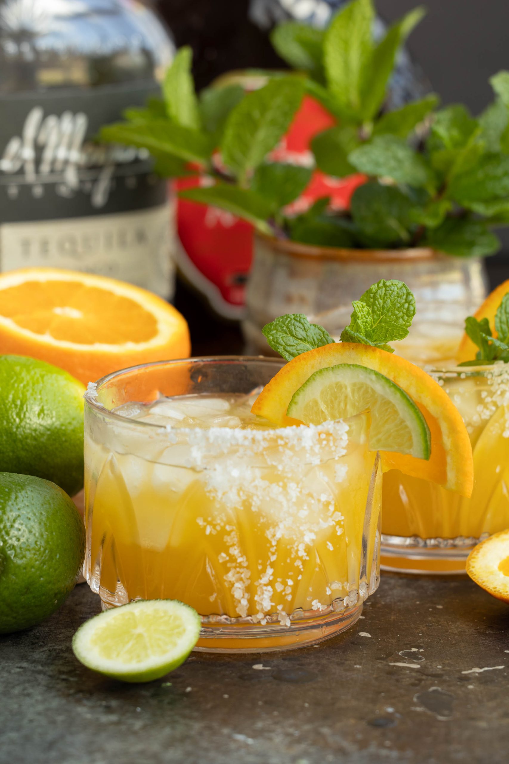 Two citrus cocktails garnished with orange slices and mint, accompanied by fresh fruit and a bottle in the background.