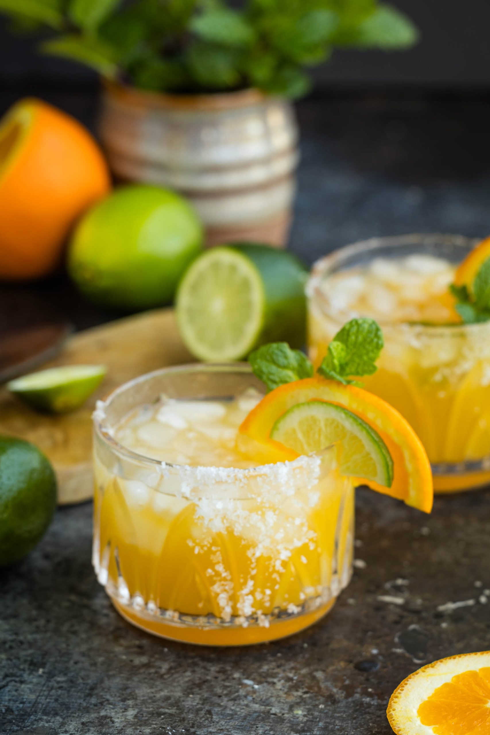 Two glasses of Italian Amaretto Margarita  garnished with lime and orange slices on a dark table, with fresh fruits and mint in the background.