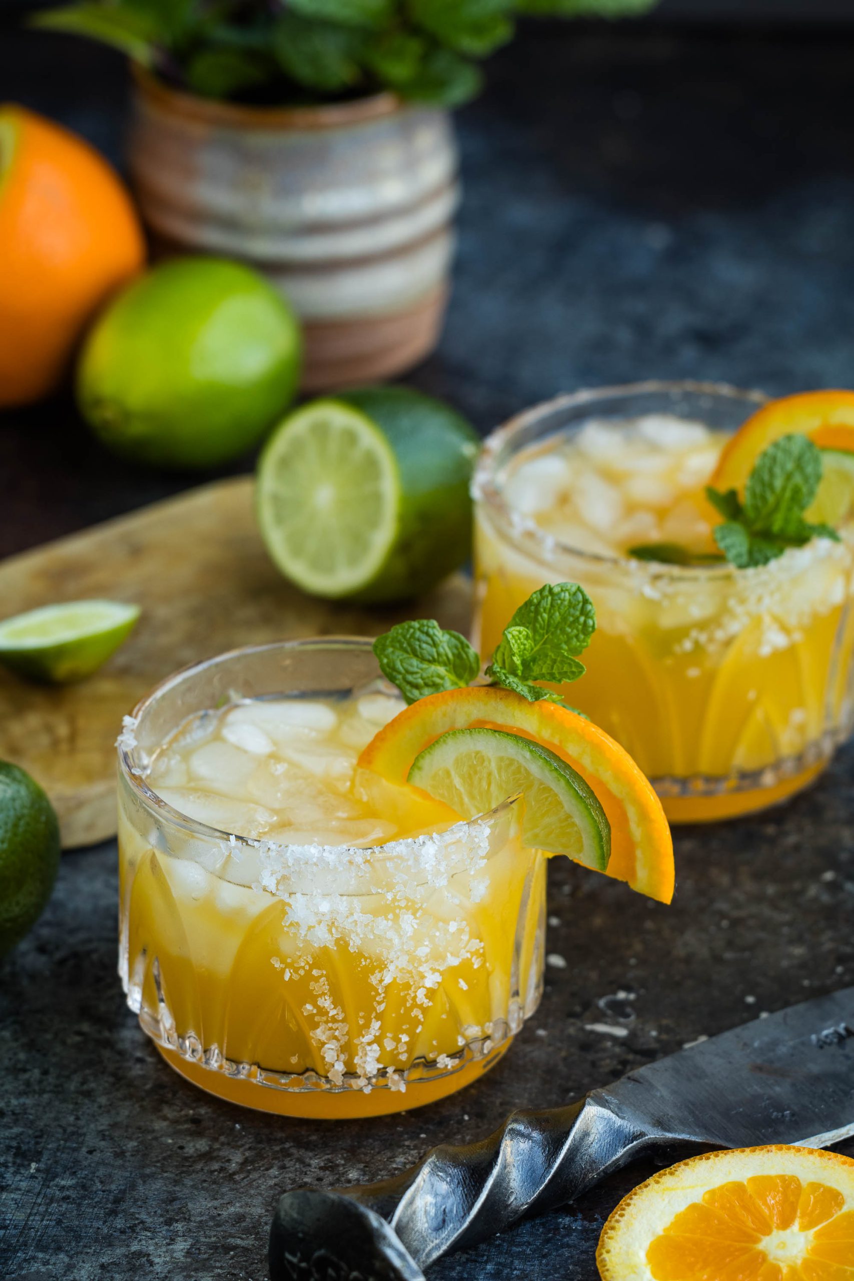 Two glasses of Italian Amaretto Margarita  garnished with lime, orange slices, and mint leaves, with a rustic background.